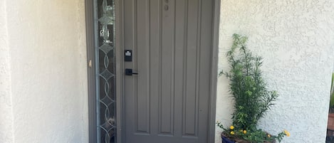 Una puerta gris con una placa de número, una planta en maceta con flores amarillas y un felpudo con la palabra 'hello'.