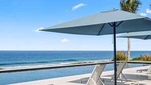 Ein Poolbereich mit Liegestühlen und einem großen Sonnenschirm mit Blick auf einen Strand und den Ozean.