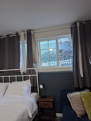 A bedroom with a bed, bedside table, lamp, and a view of a rocky garden through a window. -  Adorable Queen Bed Oakland Zoo with Private space and cool AC (Oakland)