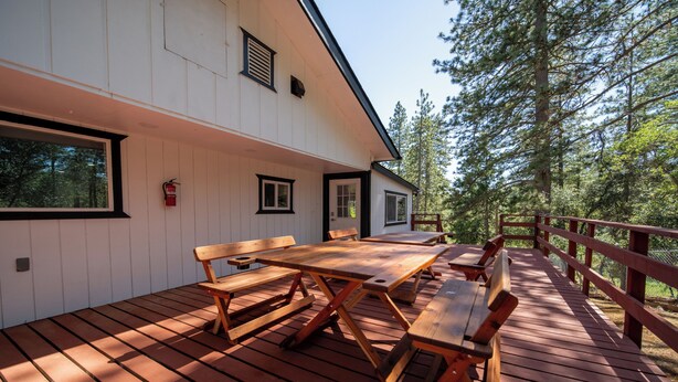 Une terrasse en bois avec une table et des bancs, un bâtiment blanc doté de fenêtres et d’une porte, et une zone boisée en arrière-plan.