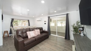 Un salon moderne avec un canapé en cuir marron, une table à manger avec des chaises, une porte-fenêtre coulissante et un téléviseur à écran plat fixé au mur.