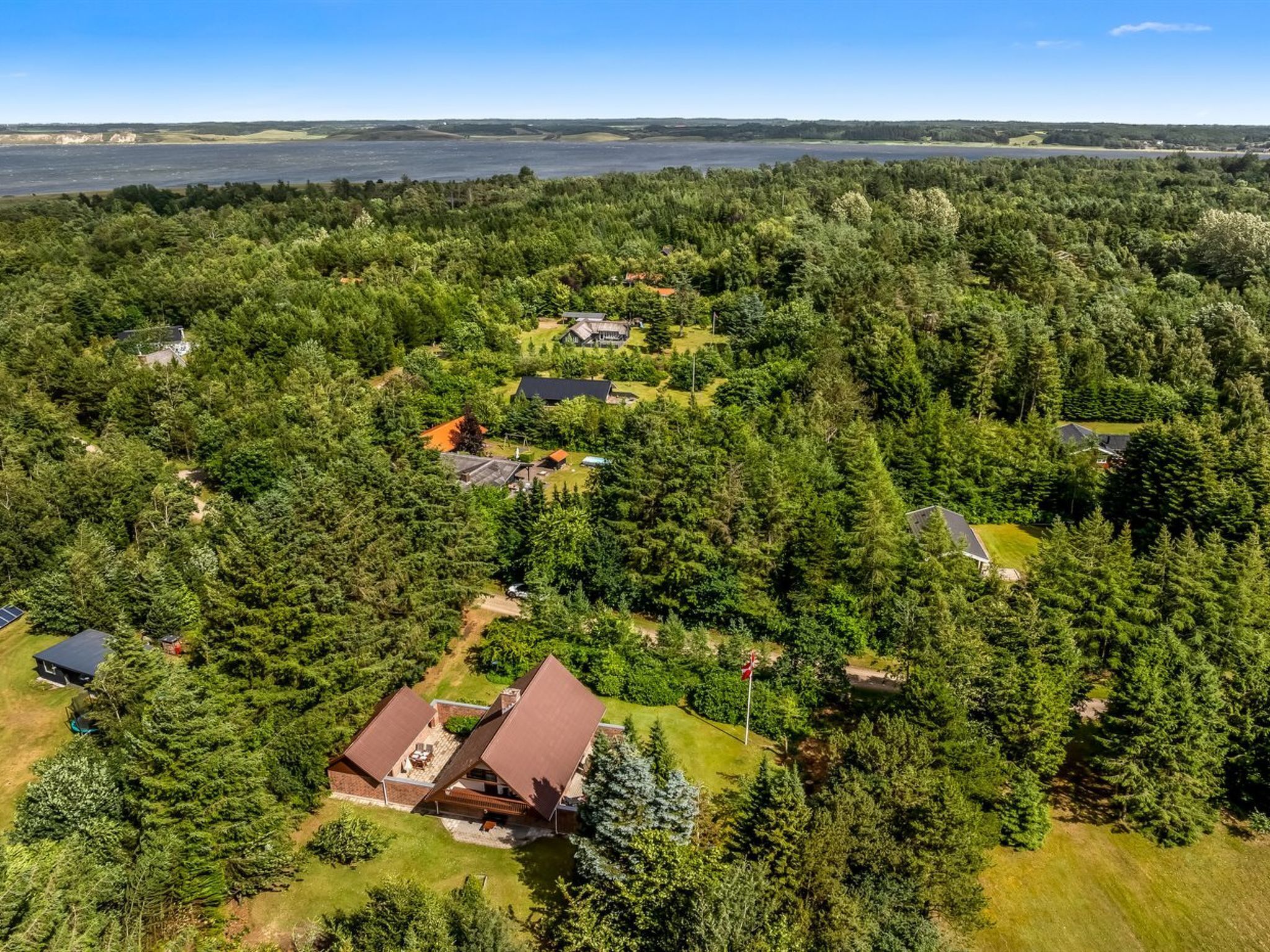 Oversigtsbillede af et boligområde med flere huse omgivet af tæt skov, en golfbane og et vandområde i det fjerne.