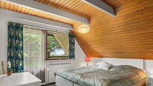 Una camera da letto con soffitto spiovente, un letto con copripiumino trapuntato, una lampada da comodino e vista attraverso una finestra con tende.