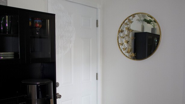 Una habitación con una puerta blanca, un mueble negro, una cafetera, un tapete pequeño y un espejo redondo con diseño floral.