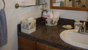Un baño con encimera de granito, un lavabo, un espejo, un perchero para toallas, una planta y una caja decorativa.