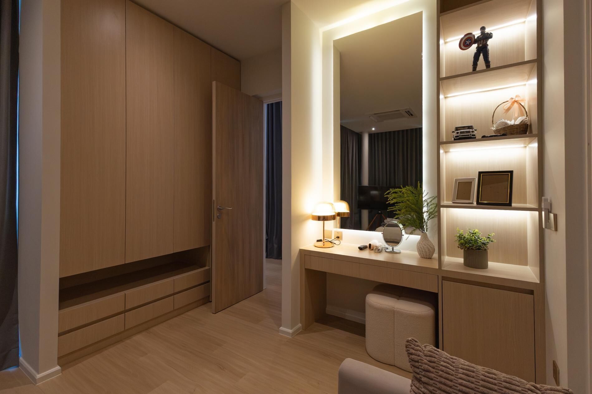 A modern hotel room with a wooden wardrobe, a vanity with a mirror, a desk with a lamp, and a small seating area.