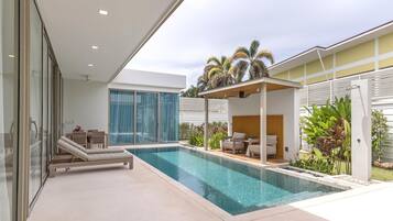 Un espace piscine moderne doté d’une pièce fermée par des baies vitrées, d’un coin salon couvert et d’une vue sur des palmiers.