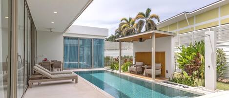 A modern pool area with a glass-enclosed room, a covered seating area, and a view of palm trees.