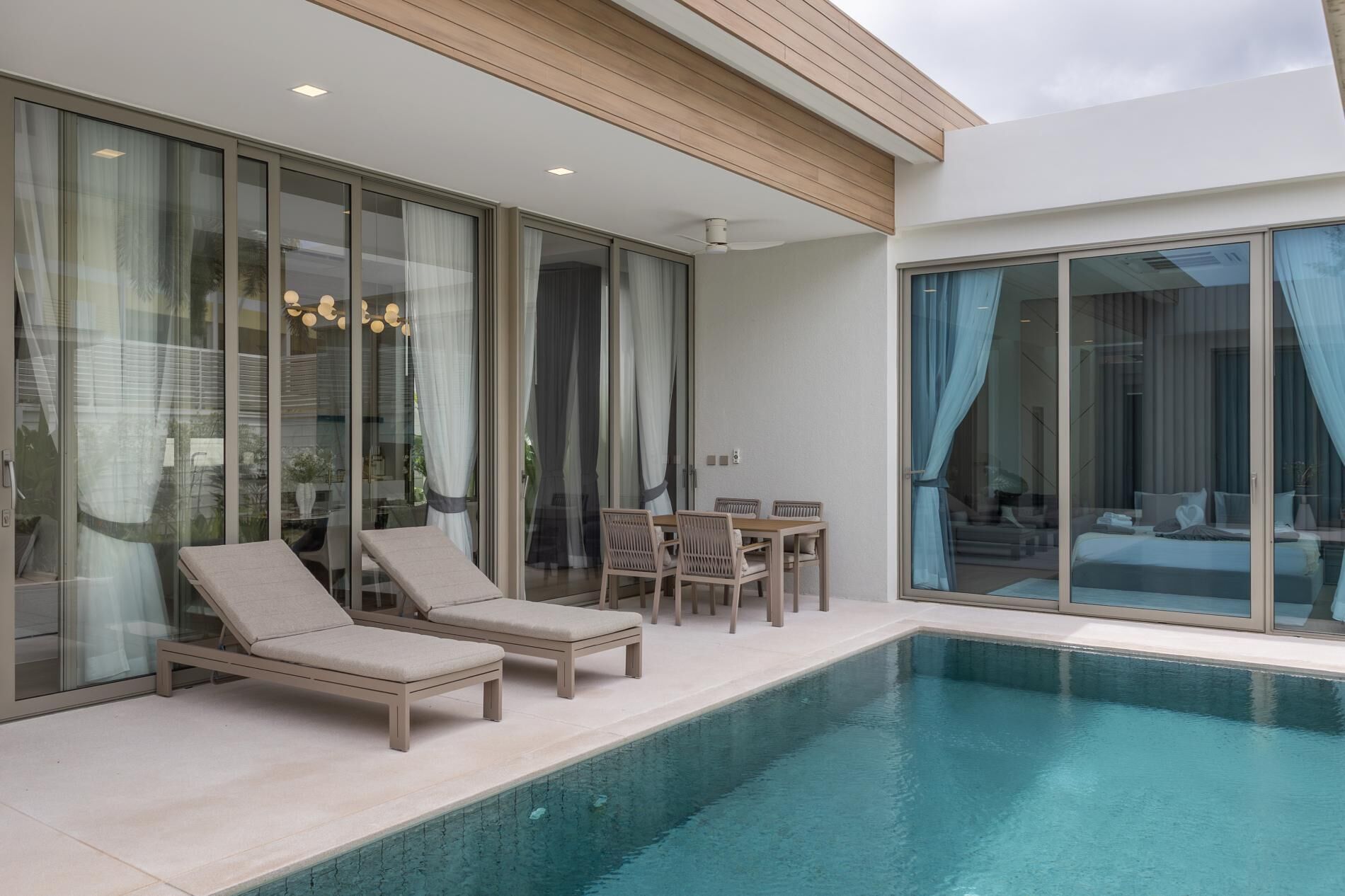 A modern pool area with lounge chairs and a dining table, large sliding glass doors, and a view of the interior.