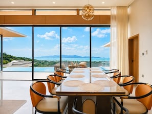 A modern dining area with a panoramic view of a coastal landscape, including an infinity pool and distant mountains.