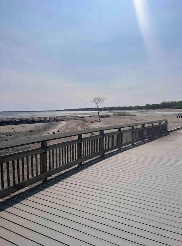 A wooden boardwalk leading to a beach with a single tree and a distant shoreline.