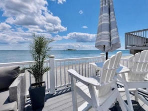 A deck with white chairs and a striped umbrella, overlooking a calm sea and a clear sky.