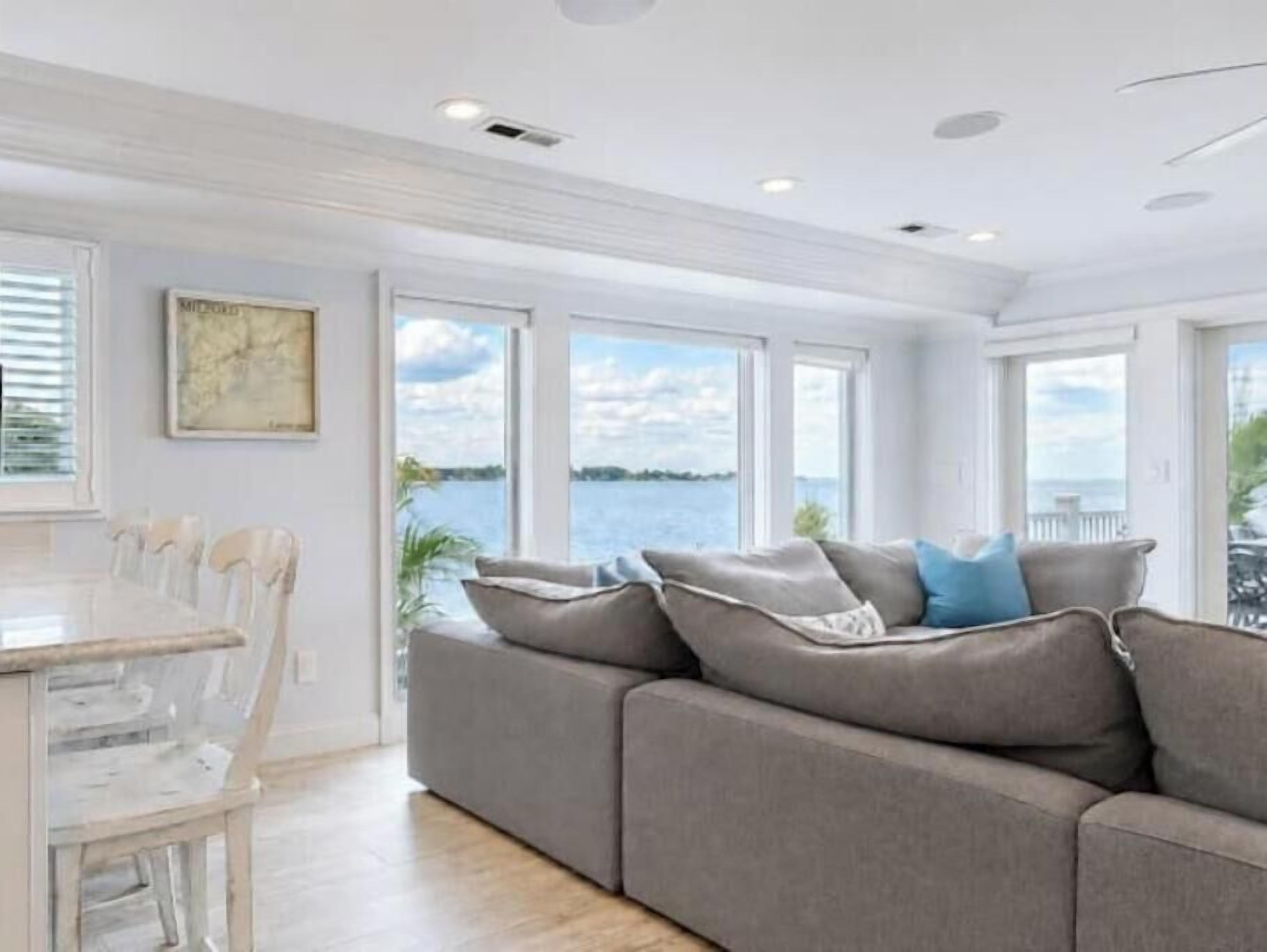 A modern living room with a gray sofa, a dining table with chairs, and large windows offering a view of the water.