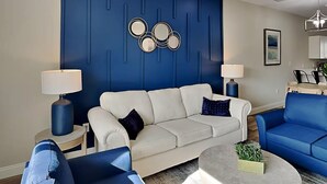 A modern living room with a white sofa, blue accent chairs, a round coffee table, and a blue accent wall with decorative mirrors. - Beach Bliss - Hot Tub, Fenced Yard 3 Bedrooms in Panama City Beach (Panama City Beach)