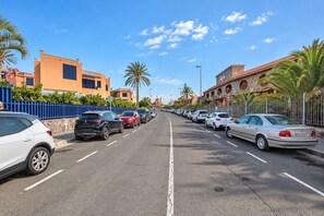 Une rue bordée de voitures garées et d’immeubles, avec notamment une clôture bleue et des palmiers.