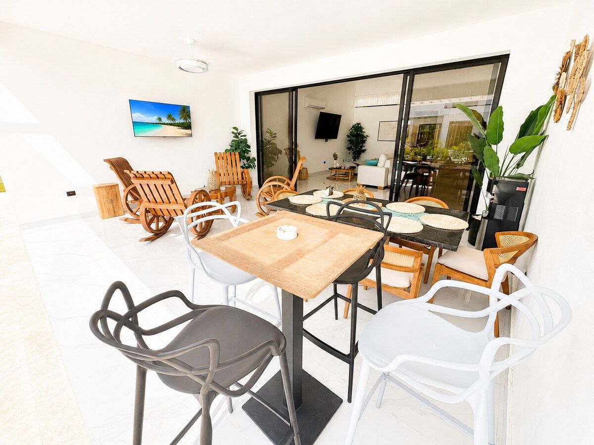 Un coin repas avec une table et des chaises en bois, un téléviseur fixé au mur et une vue sur l’extérieur.