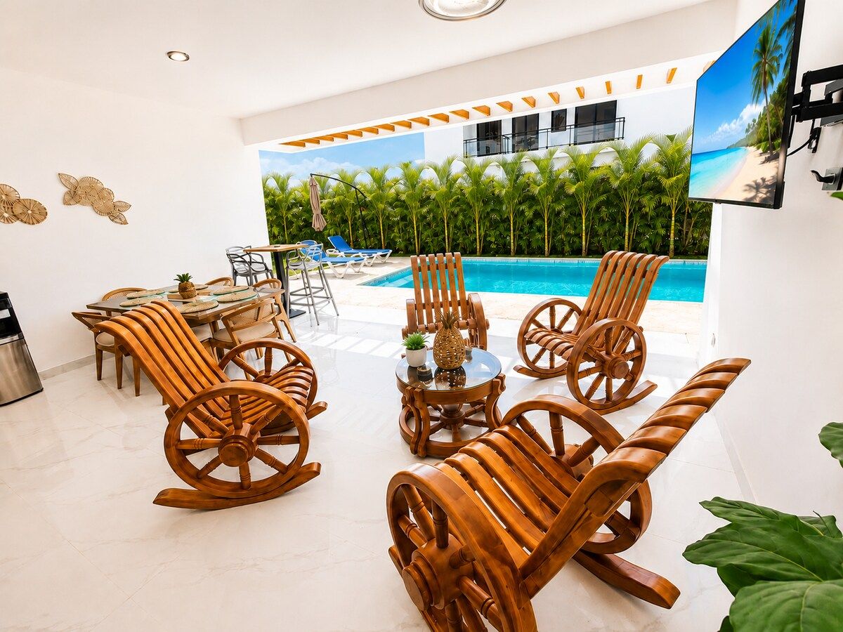 A poolside area with wooden rocking chairs, a round table, and a view of a beach through a large window.