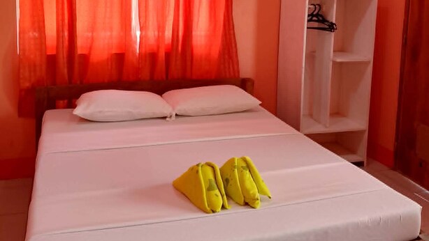 Una cama individual con ropa de cama y almohadas blancas, cabecera de madera, cortina ligera roja y armario blanco con una percha.