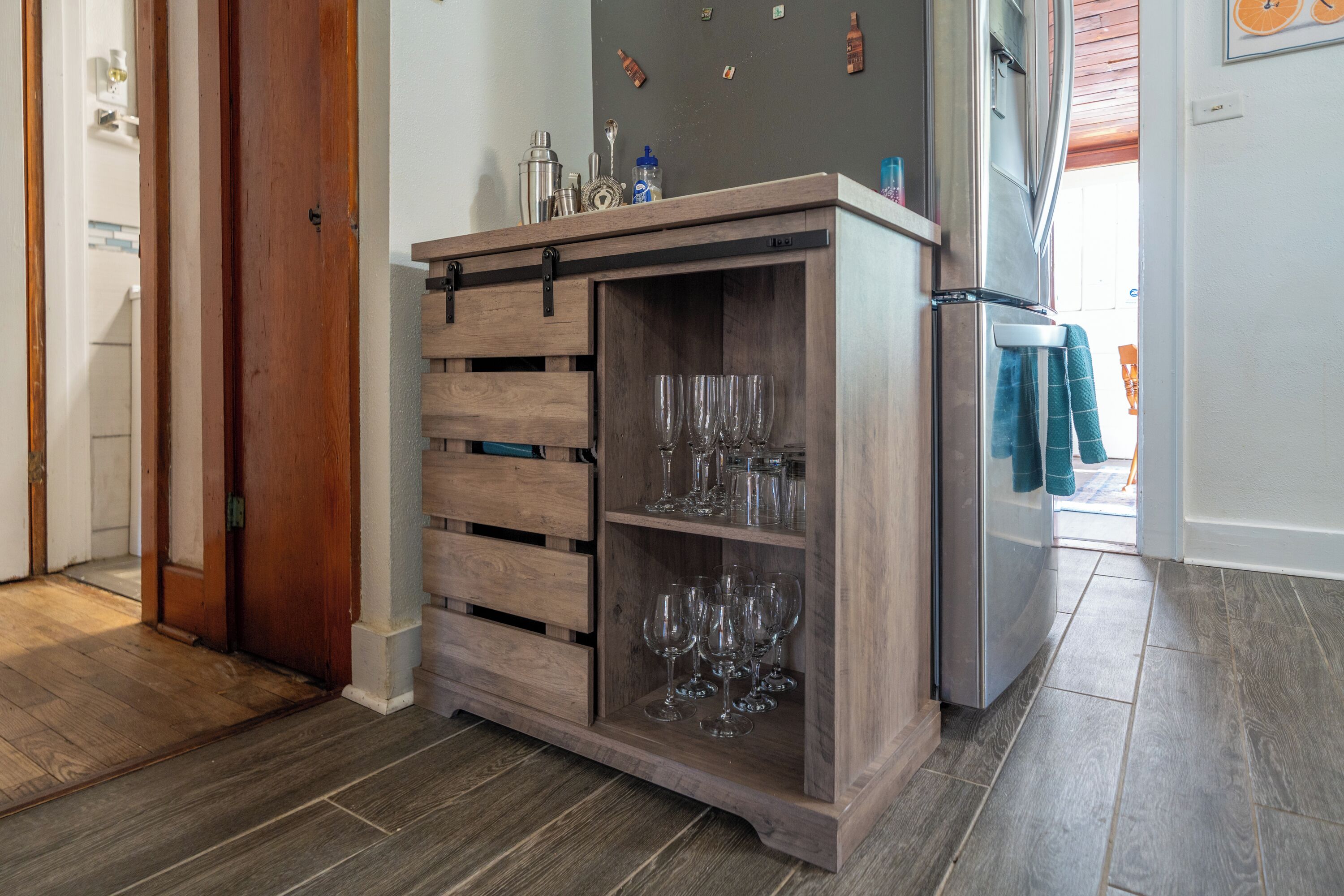 A kitchen with a wooden cabinet containing wine glasses and a stainless steel refrigerator.