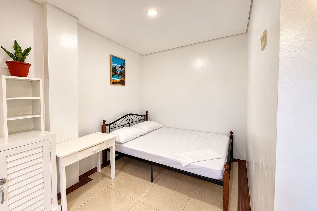 Une chambre avec un lit simple, une table de chevet, une chaise, une petite étagère et une plante en pot.