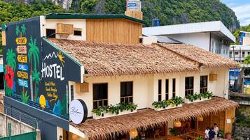 A hostel with a thatched roof, a mural advertising rooms, bar, cafe, and travel services, and a view of a mountainous landscape.