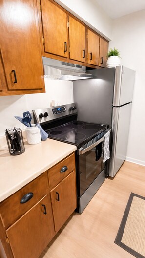 A kitchen with wooden cabinets, a stainless steel refrigerator, and a black stovetop. - Newly Remodeled + Modern Aztec Themed + 1 Bed/1 Bath Near Airport (Lansing)