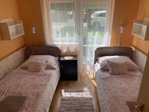 A room with two beds, a small table, and a window with a view of parked cars. - Holiday home with large garden, garden pavilion and barbecue (Zalakaros)