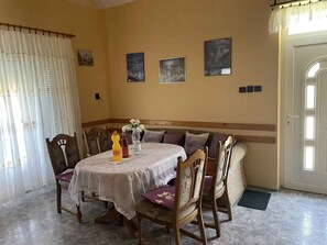 A dining area with a round table, chairs, and a bench. There are framed pictures on the wall, a door, and a window with curtains. - Holiday home with large garden, garden pavilion and barbecue (Zalakaros)
