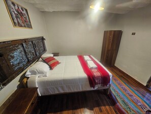 A bedroom with a wooden headboard, a bed with white bedding and a red blanket, a wooden wardrobe, and a colorful rug on the floor. - MUD HOUSE HOSTEL  (Cusco)