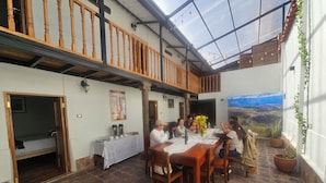 Un comedor con suelo de piedra, muebles de madera, una mesa con mantel blanco y vistas a un paisaje montañoso a través de un ventanal amplio.