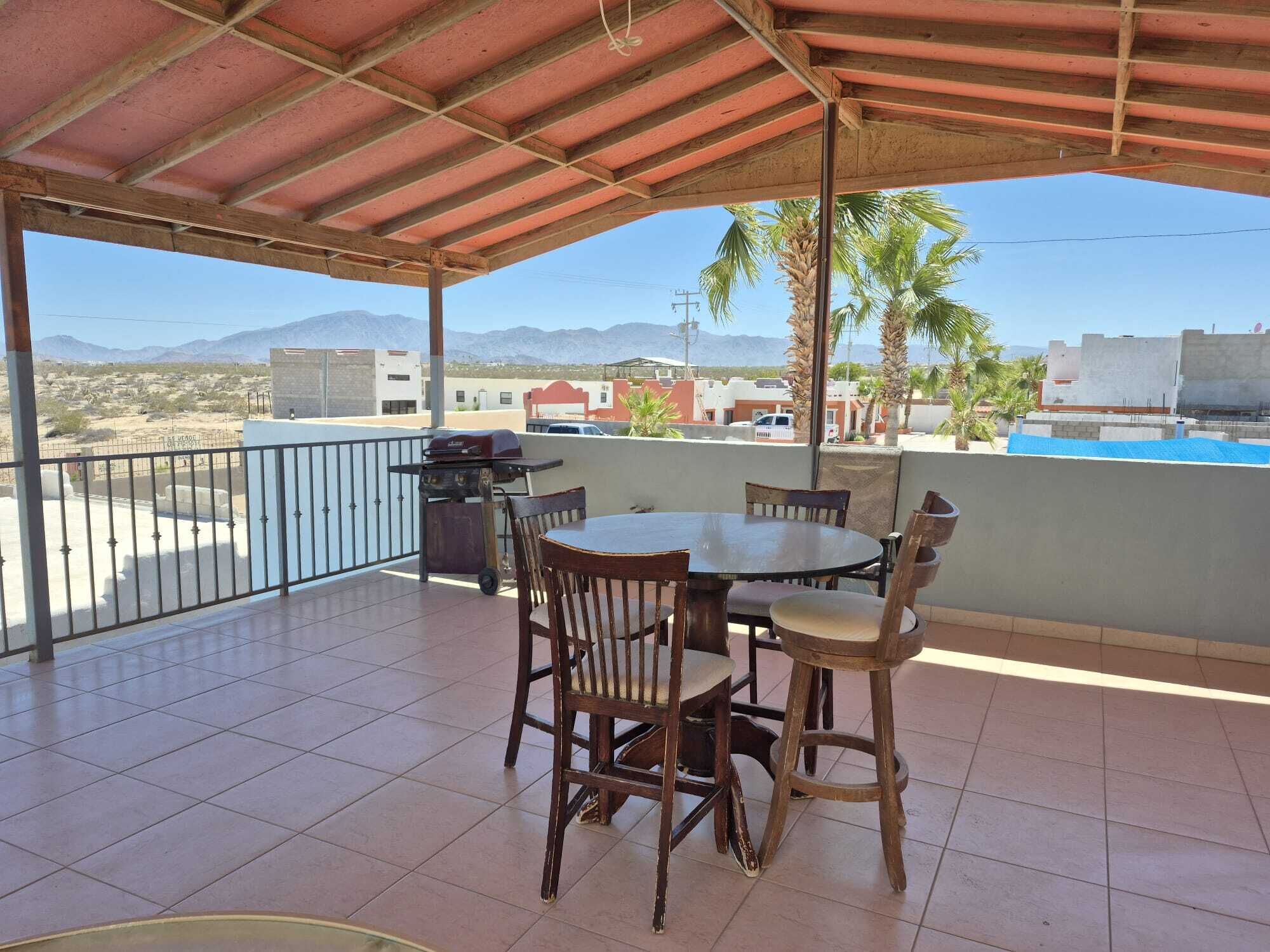An outdoor look across the covered upper patio area, featuring a wooden high-top table and chairs set, a barbecue grill, and clear blue skies over the horizon
