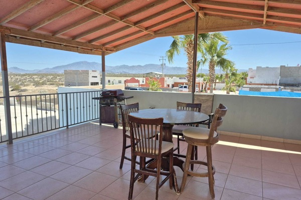 An outdoor look across the covered upper patio area, featuring a wooden high-top table and chairs set, a barbecue grill, and clear blue skies over the horizon