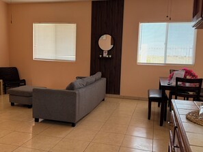 Ein Wohnzimmer mit einer Couch, einem Sessel, einem Esstisch mit Stühlen und zwei Fenstern mit Jalousien.