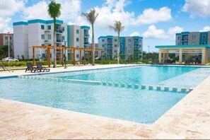 A swimming pool with lounge chairs and a pergola, surrounded by multi-story buildings with balconies. - Cozy 2BR Condo Pool Free Electricity (Punta Cana)