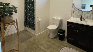Une salle de bain comprenant des toilettes, un lavabo, un miroir, une petite fenêtre, une poubelle et une plante en pot.