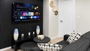 Un salon moderne avec un téléviseur à écran plat fixé à un mur foncé, un canapé gris, une petite table basse ronde et des œuvres d’art décoratives accrochées au mur.