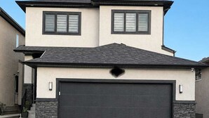 Une maison à deux étages avec une porte de garage sombre, un mur en pierre servant d’élément décoratif et un ciel dégagé.