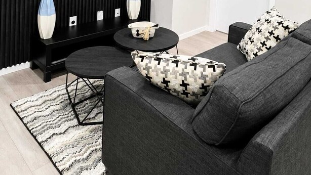 Un salon moderne avec un téléviseur à écran plat fixé au mur, un canapé noir orné de coussins à motifs, une petite table ronde et une table basse noire.