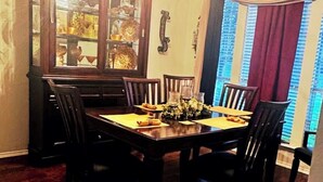 Un'area pranzo con tavolo e sedie in legno, un mobile vetrina con oggetti decorativi e vista all'esterno attraverso una finestra con tende.
