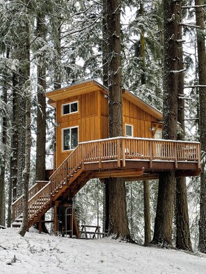Una casa en árbol de madera con una escalera, rodeada de árboles cubiertos de nieve.
