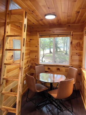 A wooden cabin interior with a round table and four chairs, a window with a view of trees, and a wooden ladder. - Mountain View Treehouse in Sutherlin with full bath and AC, 5 minutes from I-5 (Sutherlin)