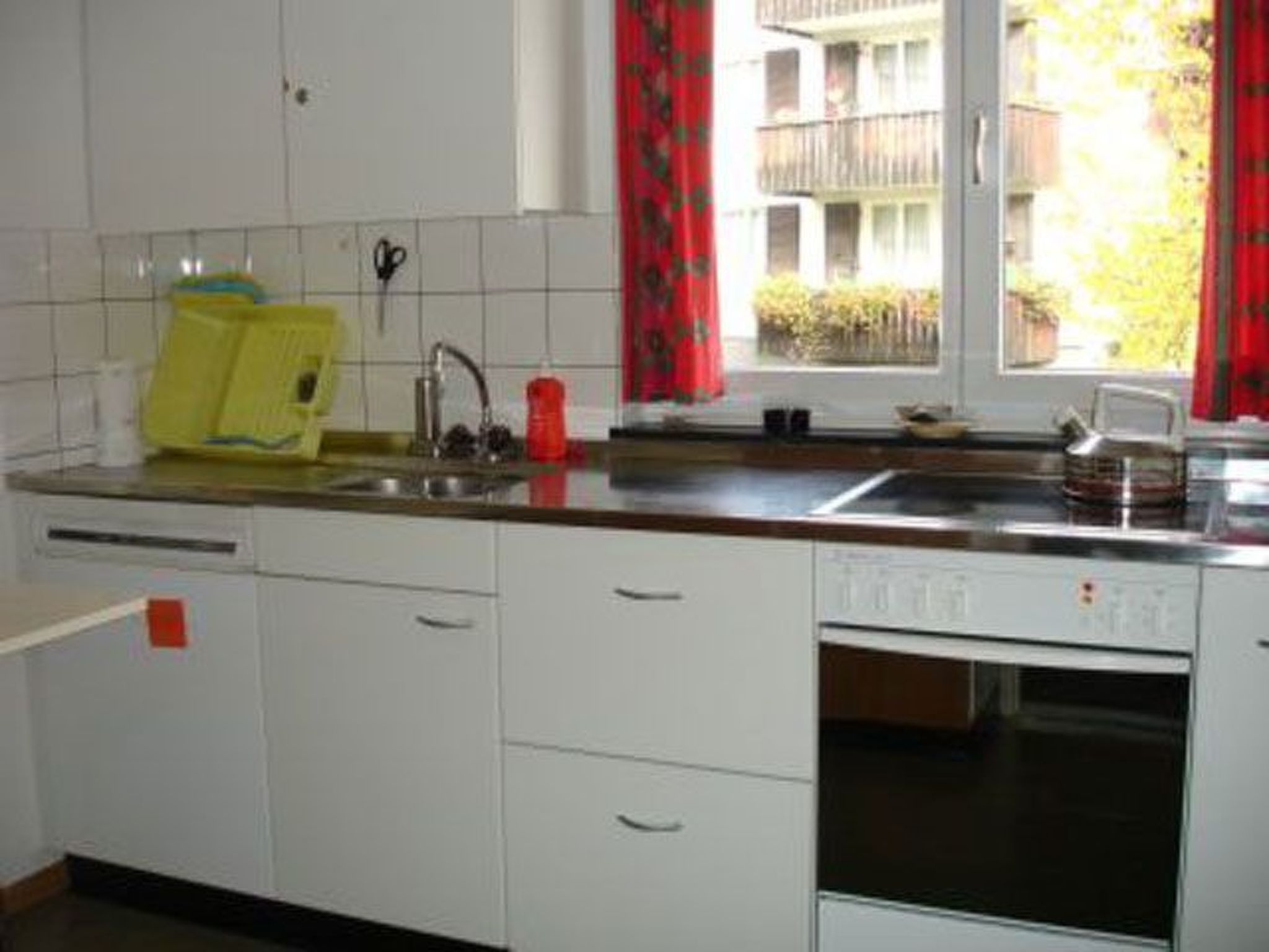 A kitchen with white cabinets, a sink, a red kettle, and a window with red curtains.