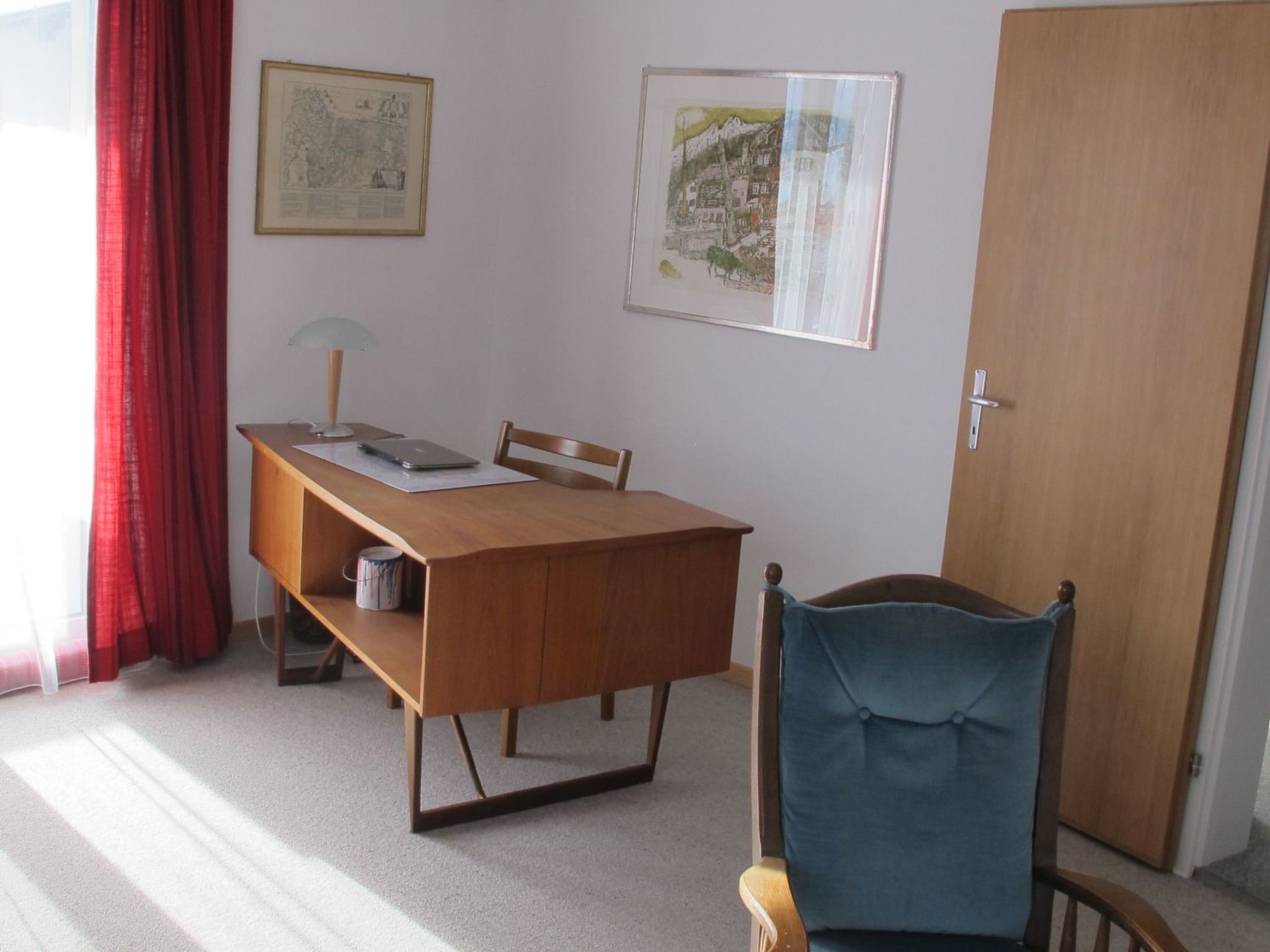Une pièce comprenant un bureau en bois, une chaise, une lampe, une porte et deux tableaux encadrés accrochés au mur.