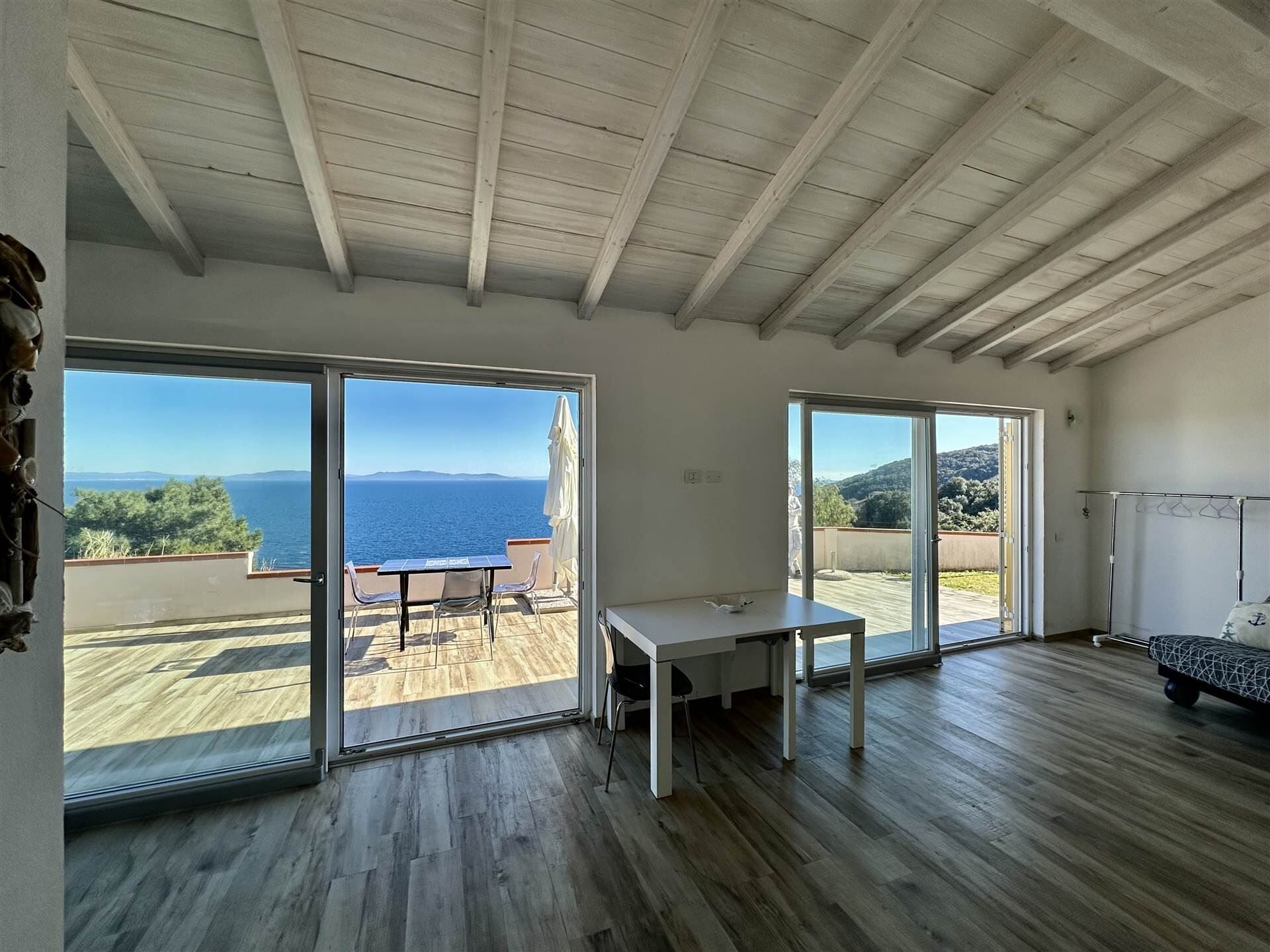 A room with a wooden ceiling, large sliding glass doors, a table, and a view of the sea.