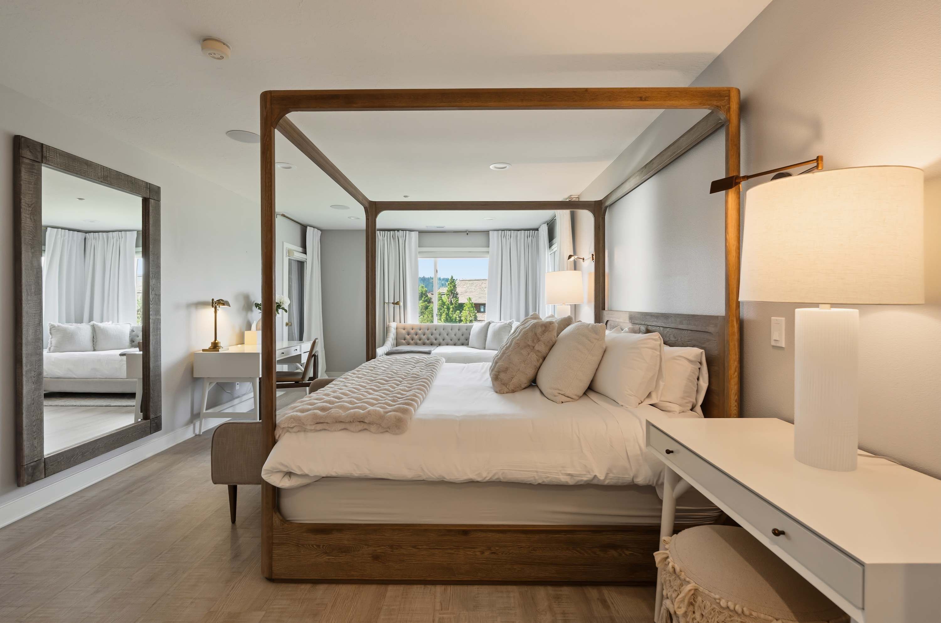A modern hotel room with a four-poster bed, a sitting area with a bench, a large mirror, and a view of the outdoors.