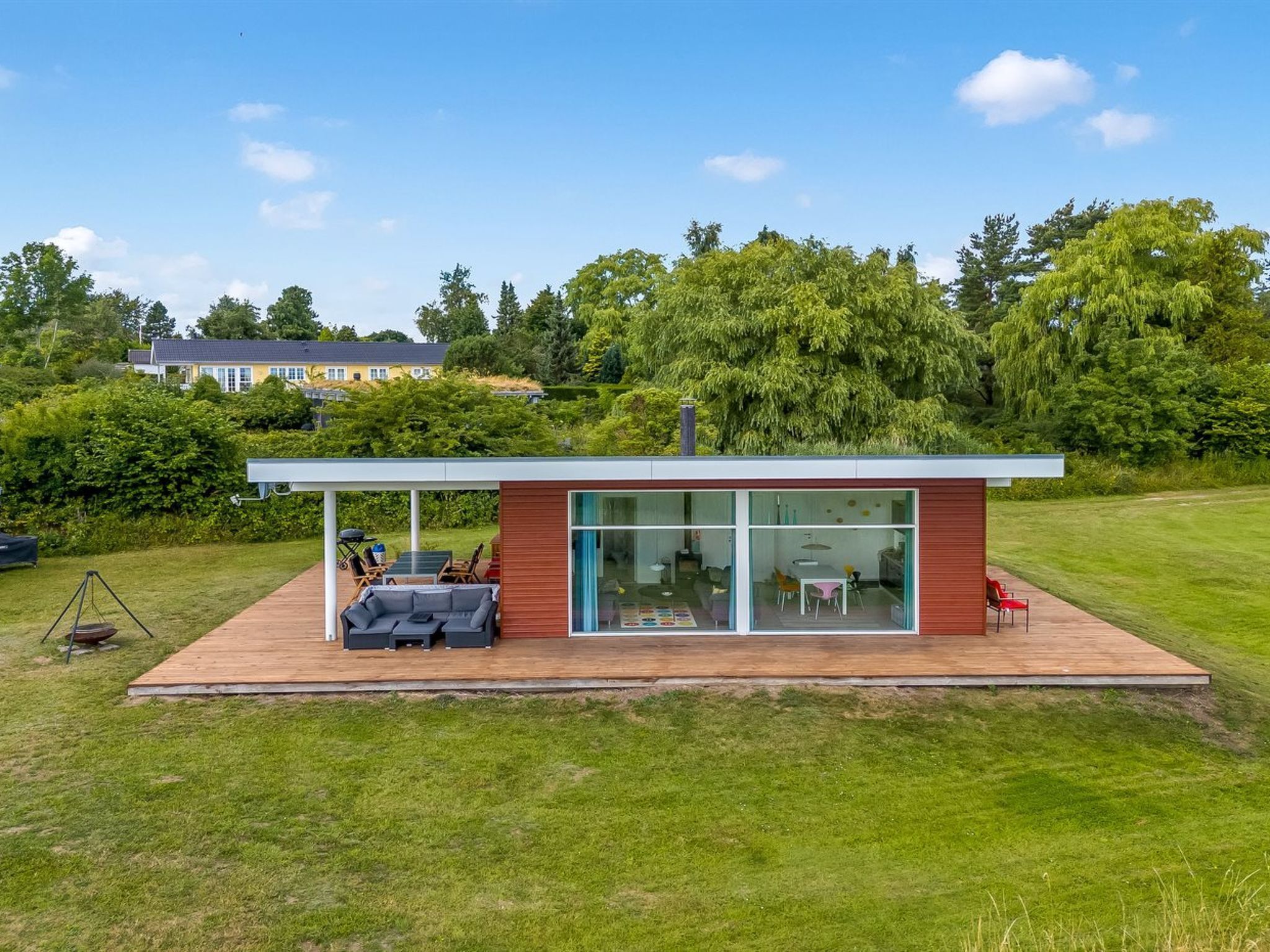 Ett modernt hus med en inglasad uteplats, utomhusmöbler och utsikt över en frodig trädgård.