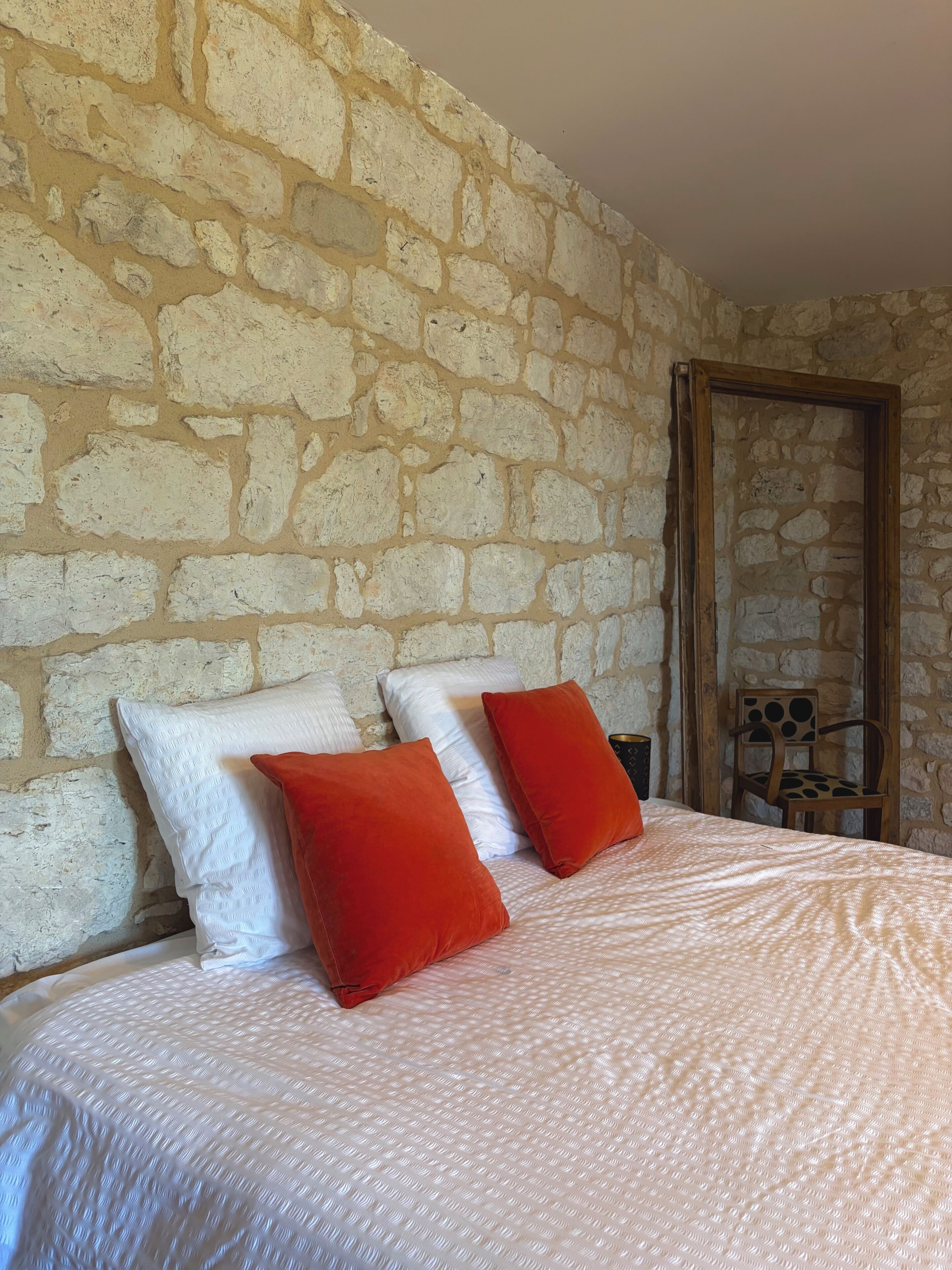 Una habitación con pared de piedra, una cama con almohadas blancas y rojas, y una silla de madera.