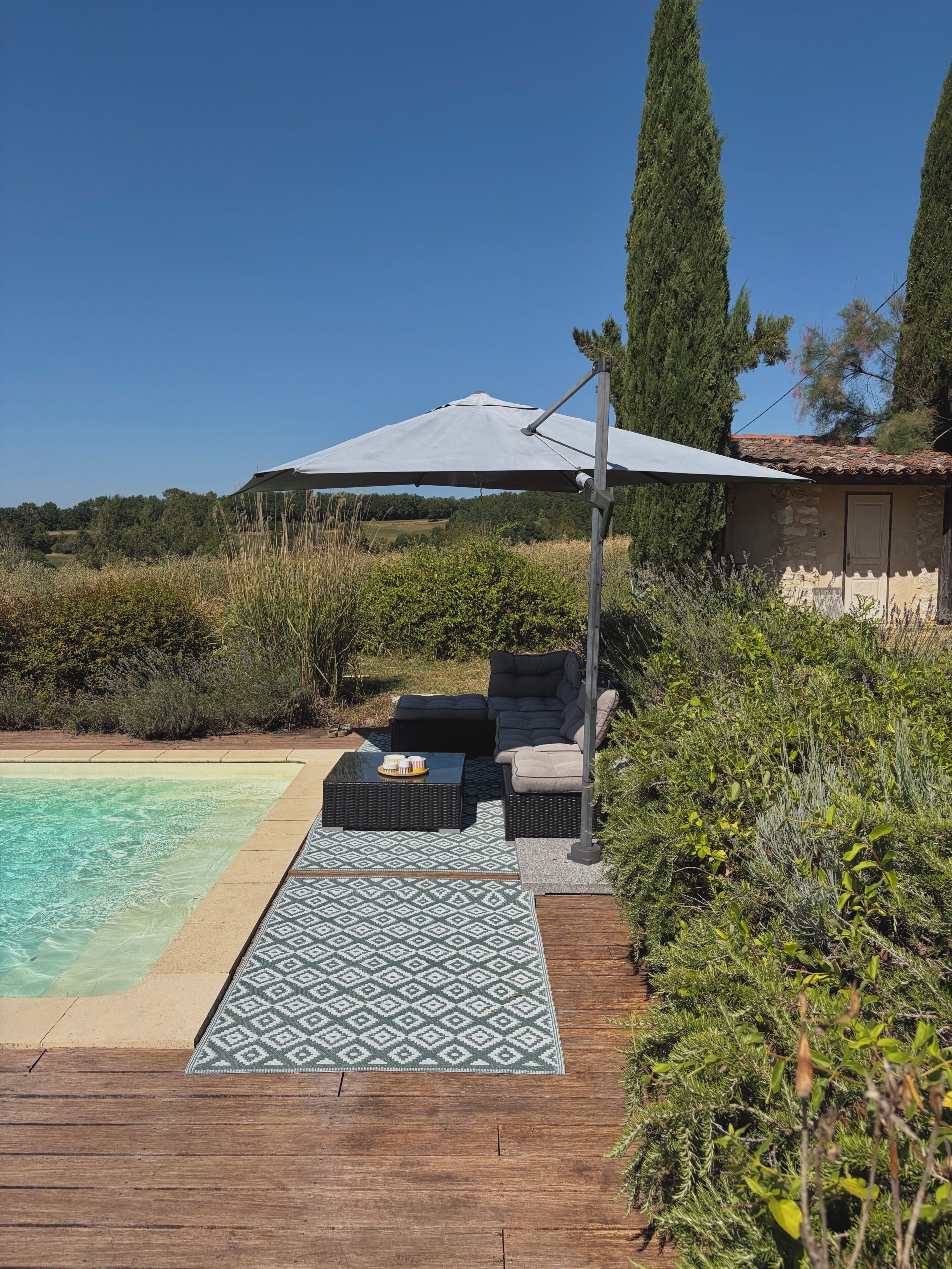 Un espace aménagé au bord de la piscine, avec un coin salon couvert, un tapis à motifs et un bâtiment en pierre en arrière-plan.