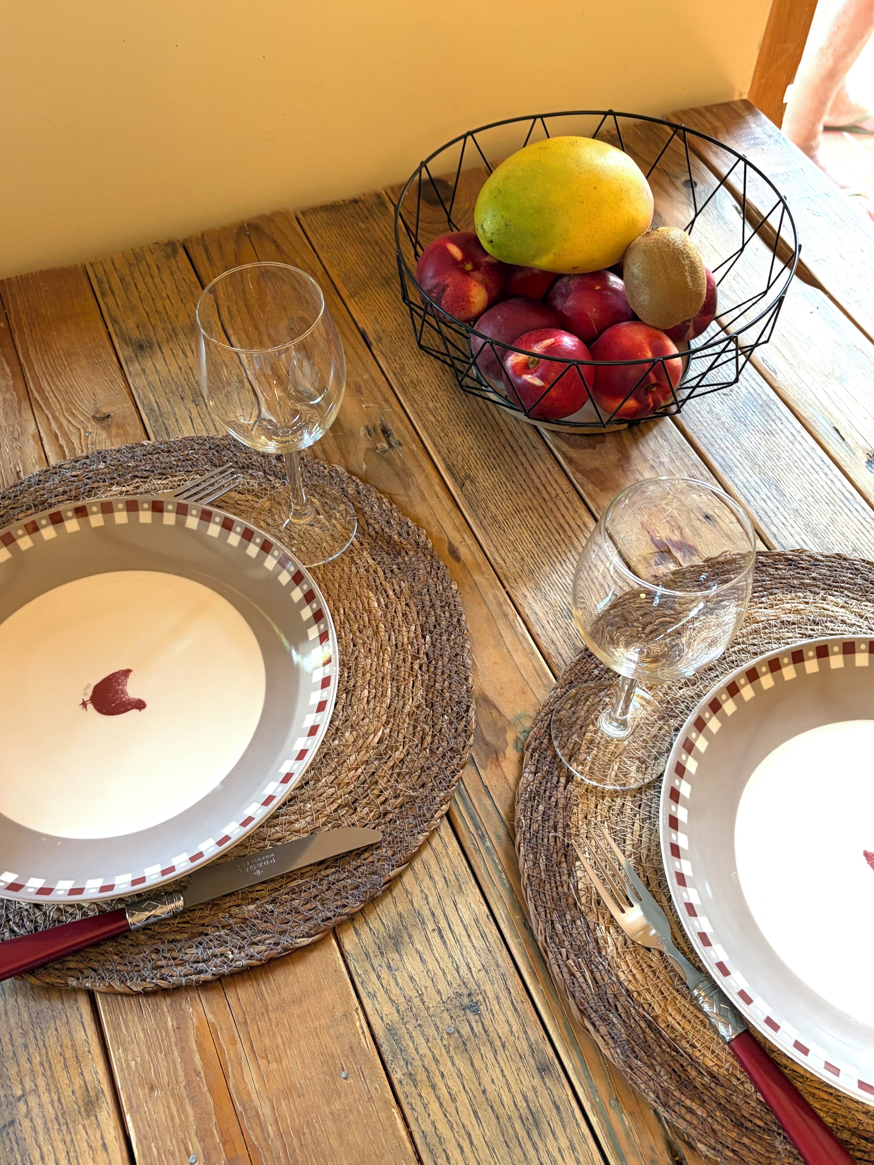 Una mesa de madera con una cesta de frutas, dos copas de vino y un plato con una flor roja.