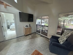 A living room with a flat-screen TV, a sofa, a wooden cabinet, and a view of a backyard through a sliding glass door.